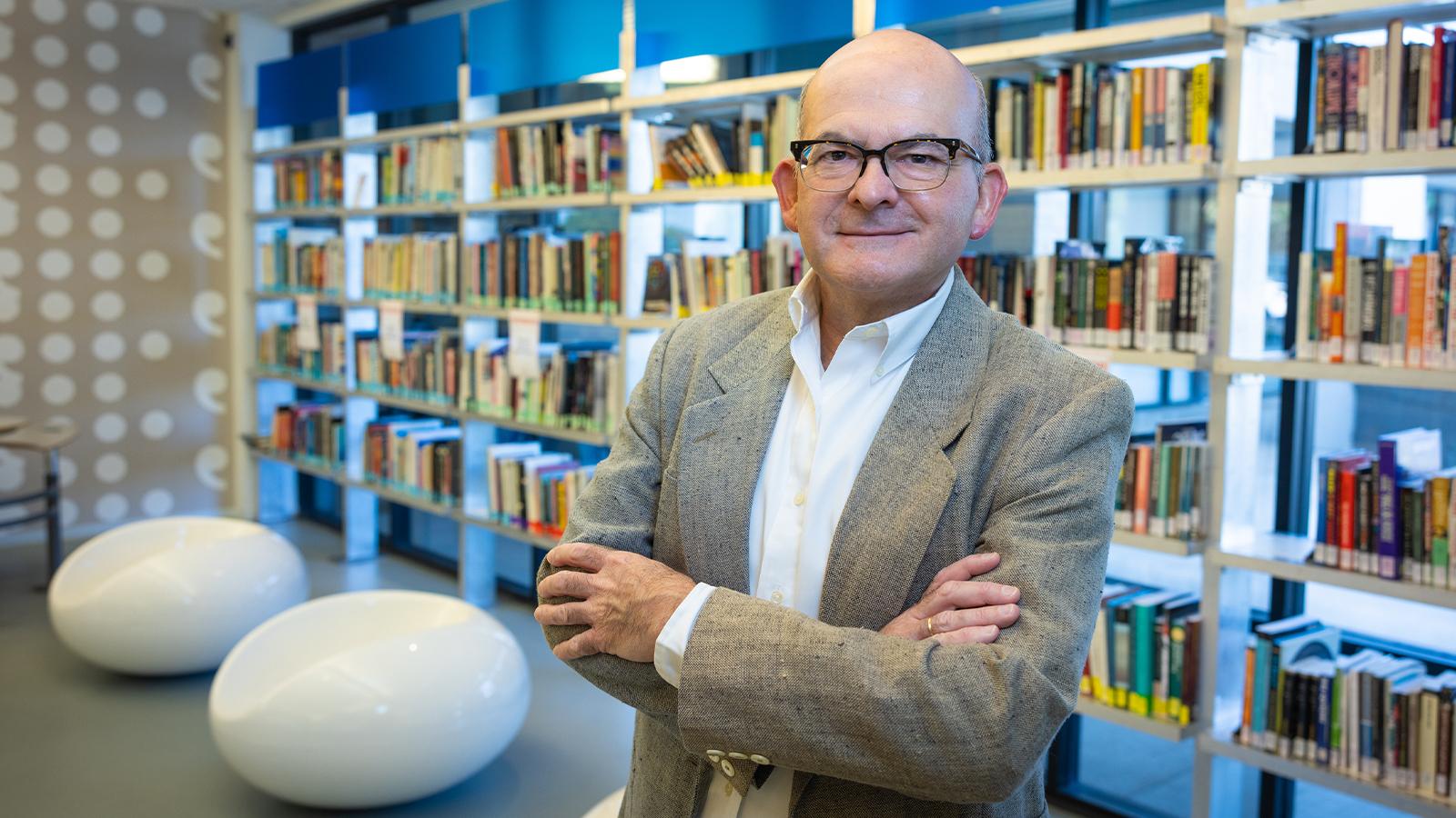 Rol和o st和s in a library with his arms crossed. He is smiling 和 wearing a suit.