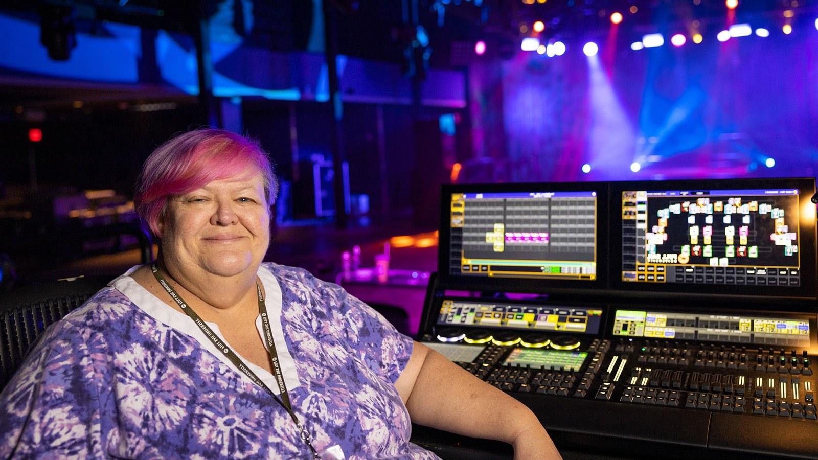 苏珊·凯莱赫坐在灯光控制台前. 她身后的舞台被紫色、蓝色和白色的灯光照亮.