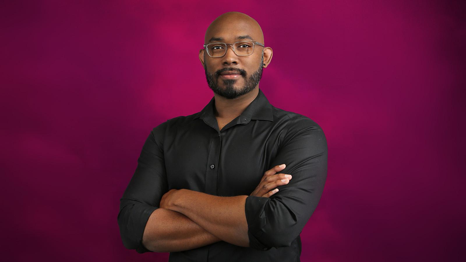 Gregg is wearing a black shirt and rectangular glasses. He is standing in front of a pink background with his arms crossed.