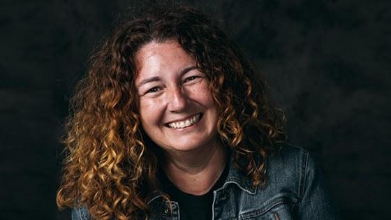 A woman with curly auburn hair smiling while wearing a black tee and denim jacket against a black backdrop.