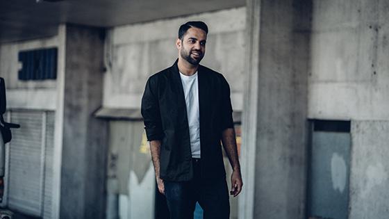 A well-dressed man in a white t-shirt and black blazer posing in front of a worn-down building.