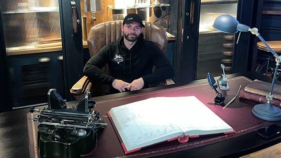 Rob Prager sits in a leather chair behind a large desk. He is smiling and wearing a black baseball cap and a black hoodie.