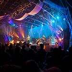 乐队在一个大音乐厅演奏音乐, dark auditorium with several bright blue 和 purple lights pointing at the stage.