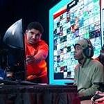 Two Florida high school students seated on stage in front of a gaming monitor with a Full Sail Armada athlete helping them to their right.