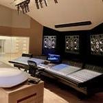 A conference table faces a wall with mixing boards and speakers; a recording area with a grand piano is in the background.