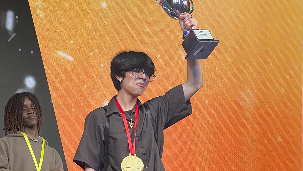 A young man on stage holding up a silver trophy over his head while wearing a large gold medal around his neck.